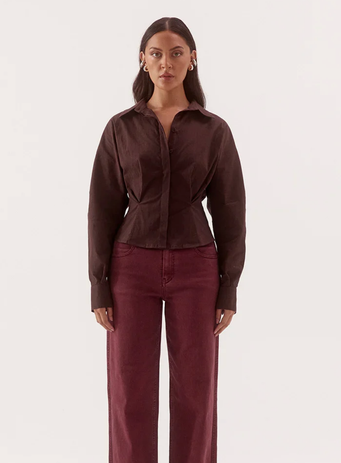 A woman modeling a dark brown long-sleeve button-up shirt paired with burgundy wide-leg jeans against a plain white studio background.