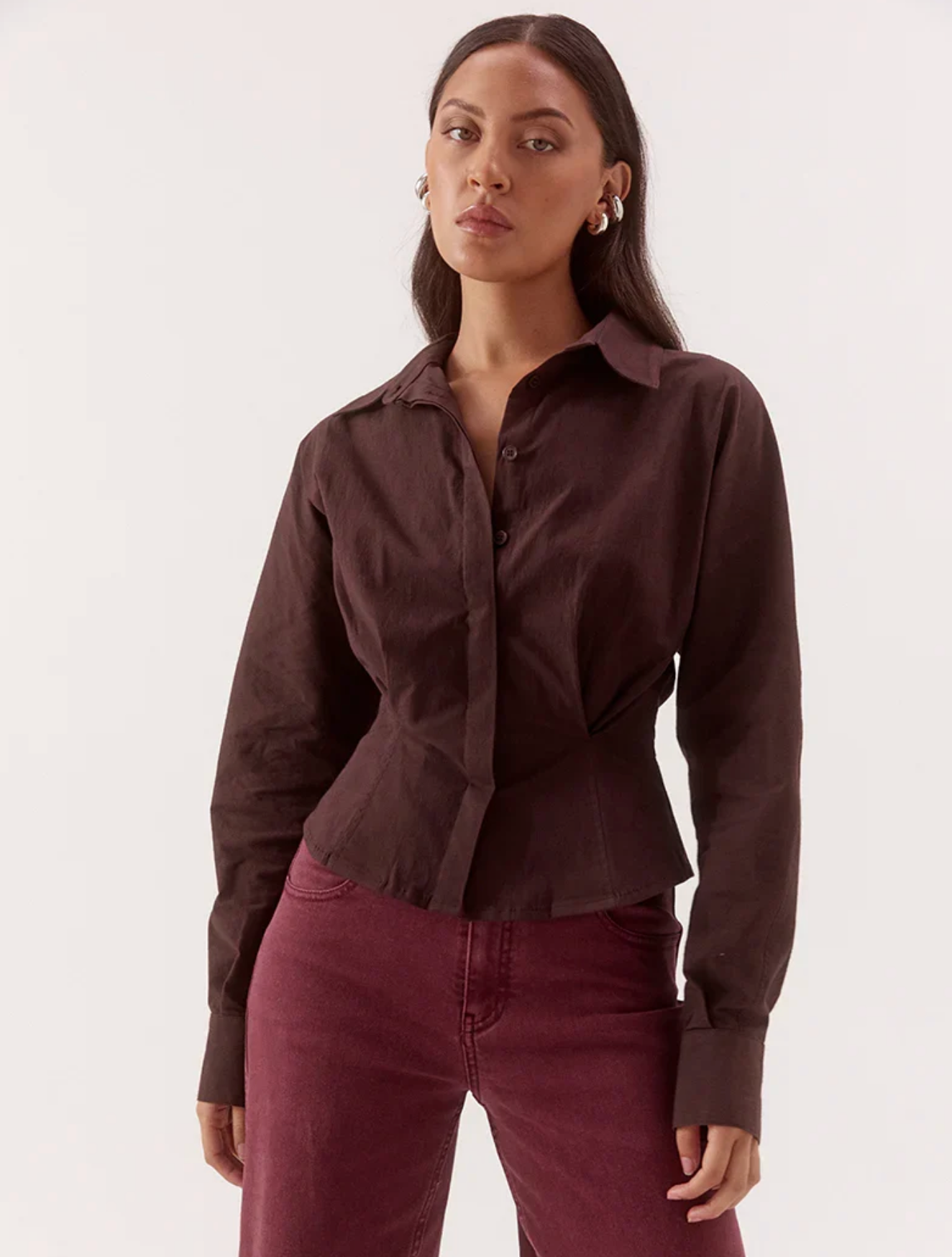 A woman modeling a dark brown long-sleeve button-up shirt with a unique gathered waist detail, paired with burgundy pants, against a plain white studio background.