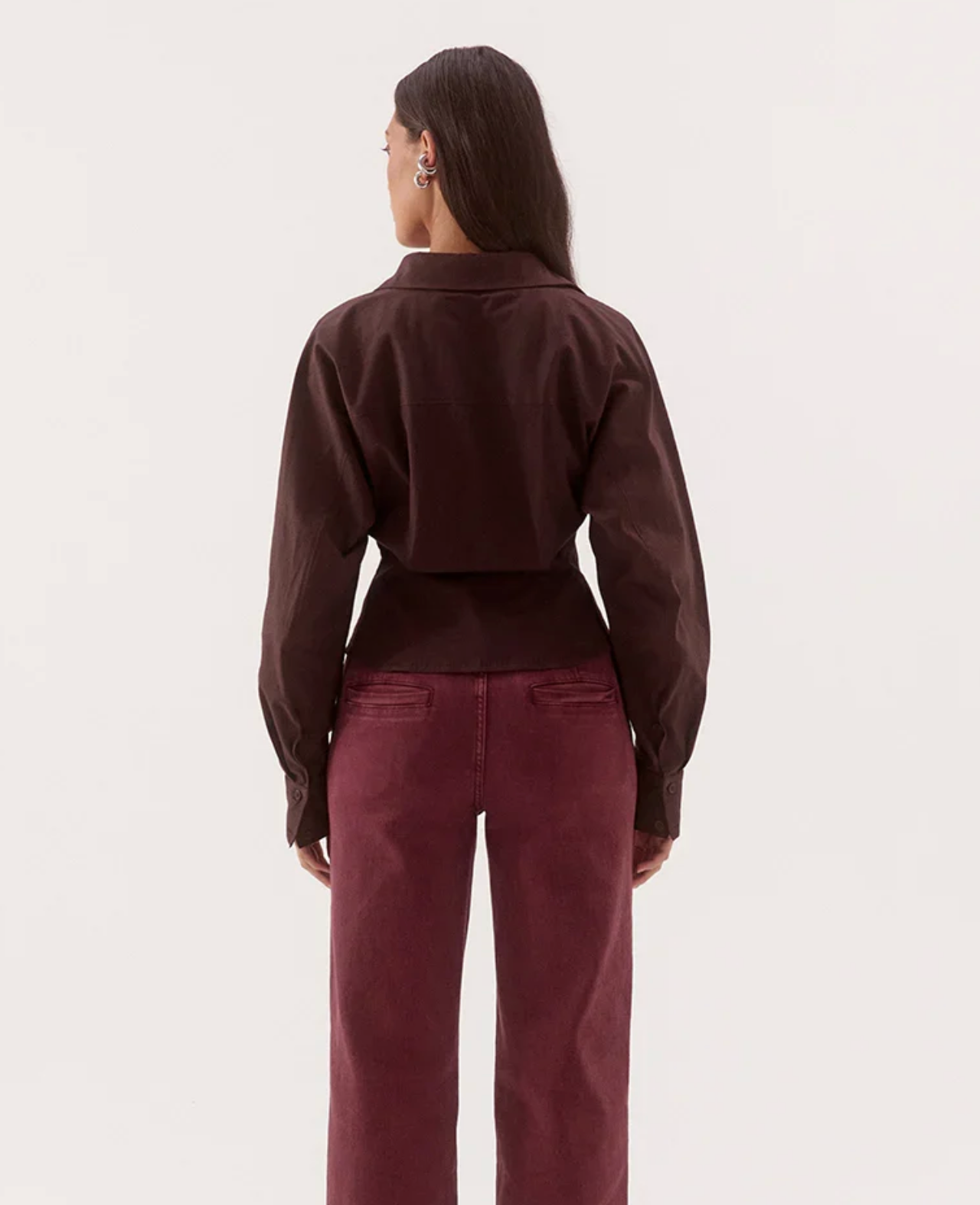 A model shown from the back wearing a dark brown cropped long-sleeve collared shirt and burgundy wide-leg pants, set against a plain off-white studio background.