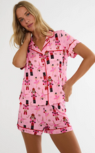 A woman modeling a pink short-sleeve pajama set with an all-over nutcracker and ballerina print, featuring a button-up top and matching shorts, against a plain white studio background.