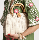 Person holding a woven handbag with a bamboo handle, wearing a green embroidered garment.