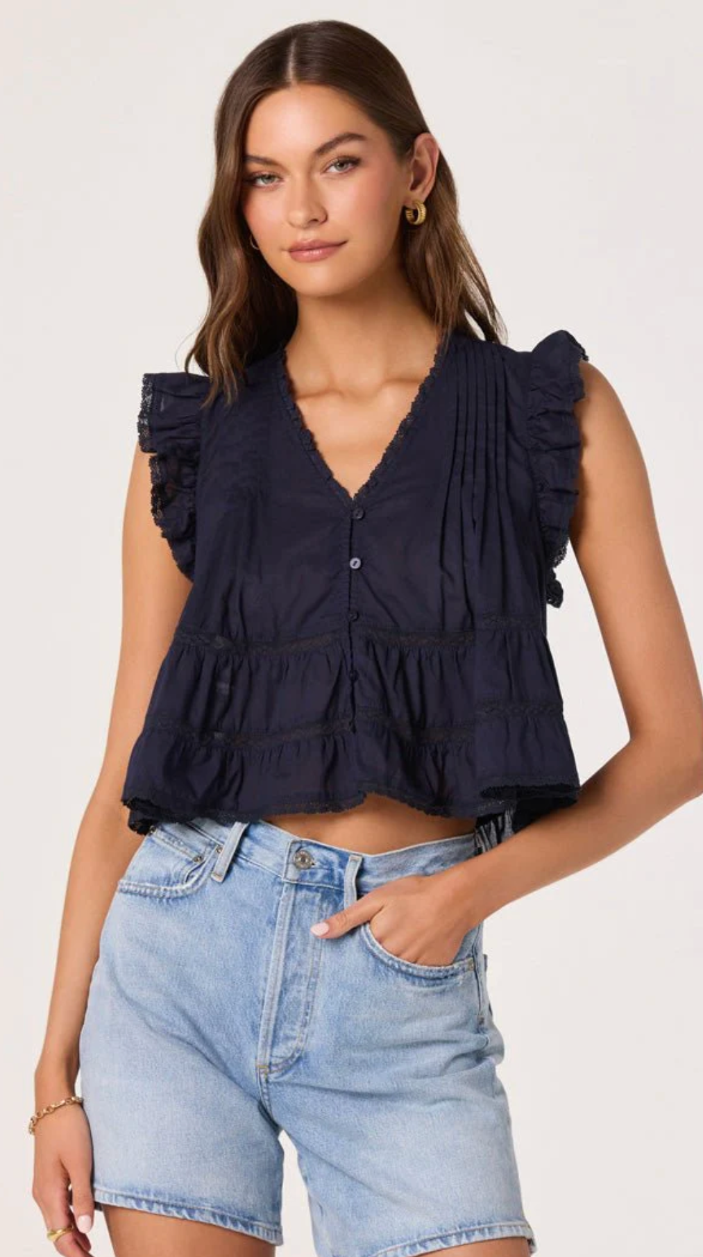 Woman wearing a navy sleeveless top with ruffled details and light blue denim shorts on a white background