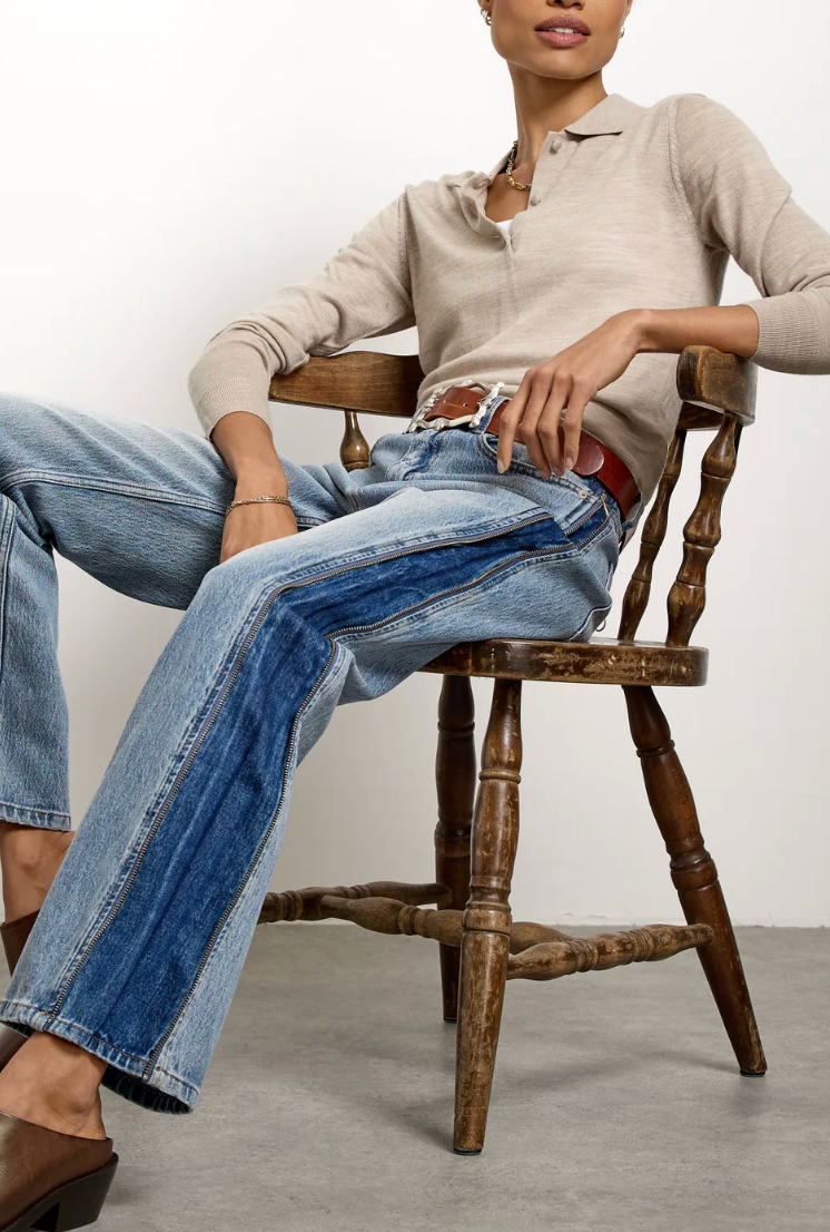 A model sits on a rustic wooden chair wearing a beige long-sleeve henley top, blue wide-leg jeans, and a red belt. The photo is taken in a clean studio setting with a white wall and gray floor.
