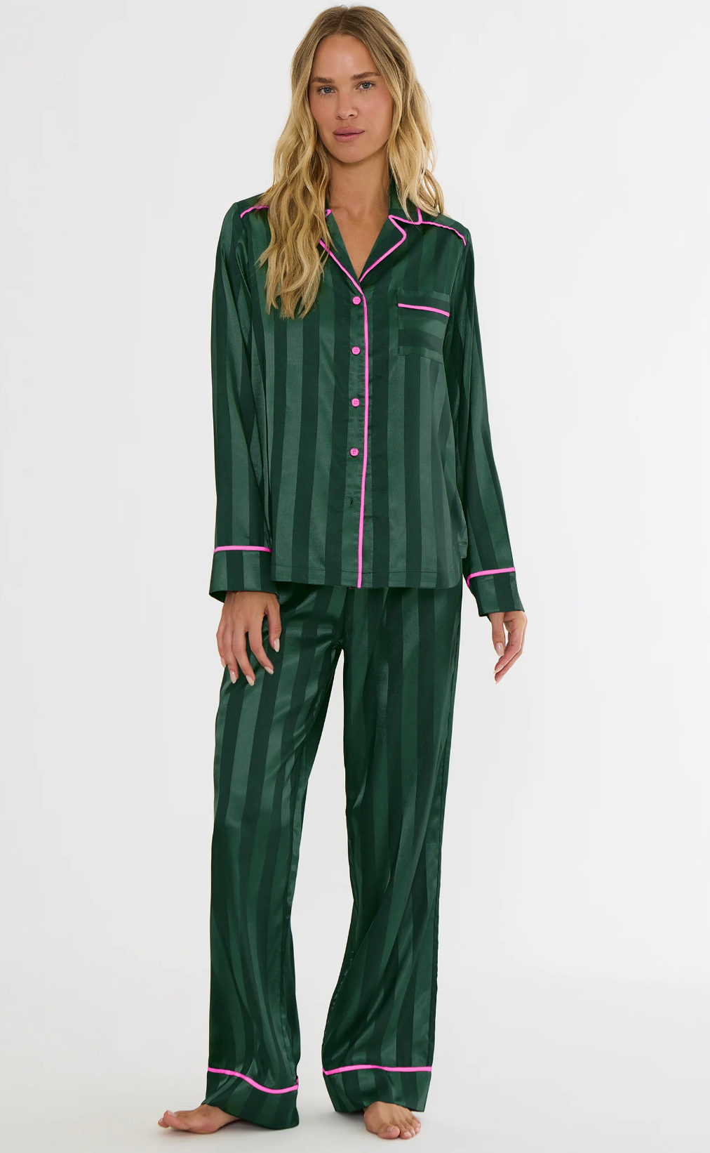 A woman modeling a two-piece green pajama set with vertical stripes and pink piping details, standing against a plain white studio background.