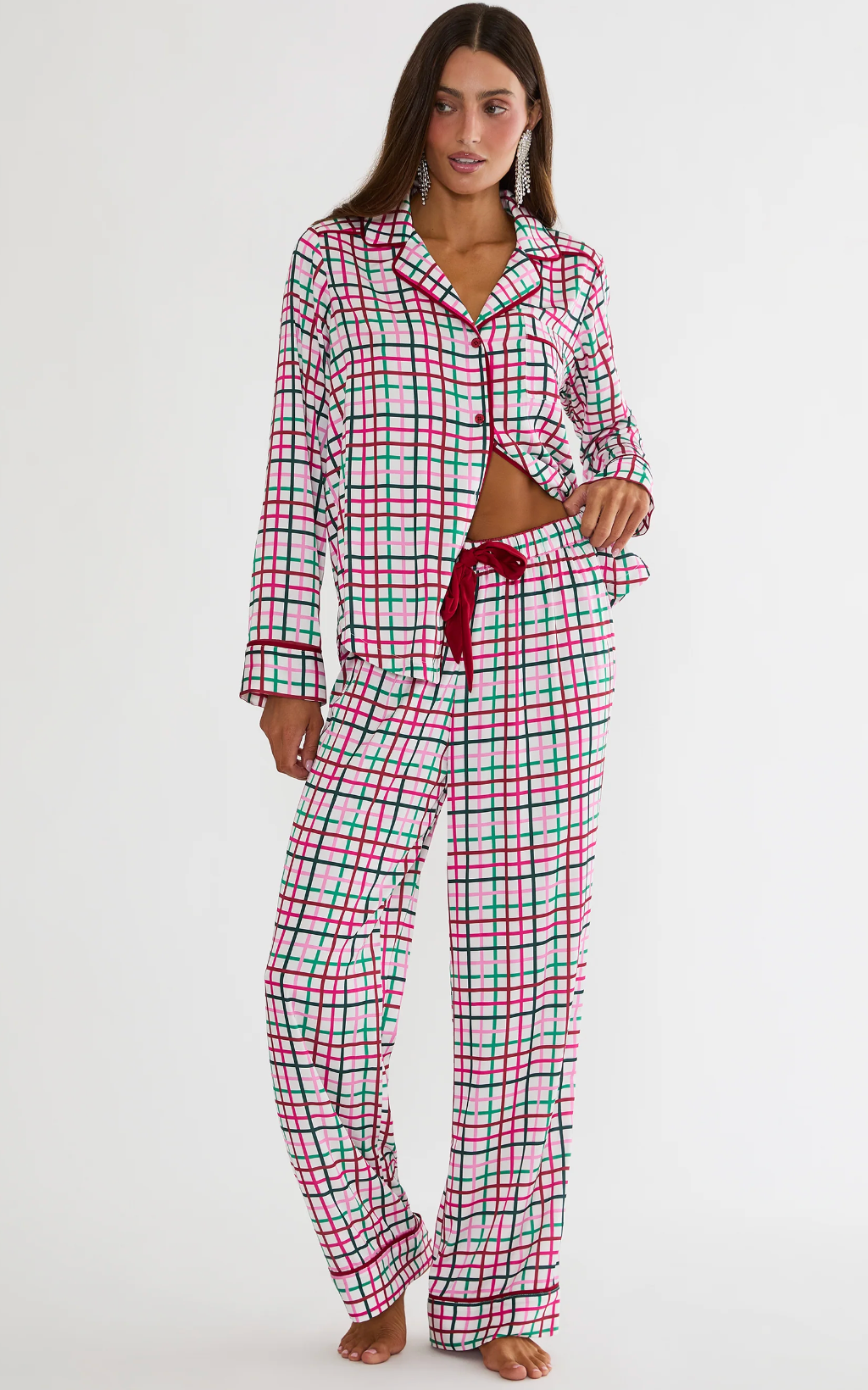 A woman modeling a two-piece pajama set with a red, green, and white plaid pattern. The set includes a long-sleeve button-up top with a red tie waist and matching wide-leg pants, photographed against a plain white studio background.