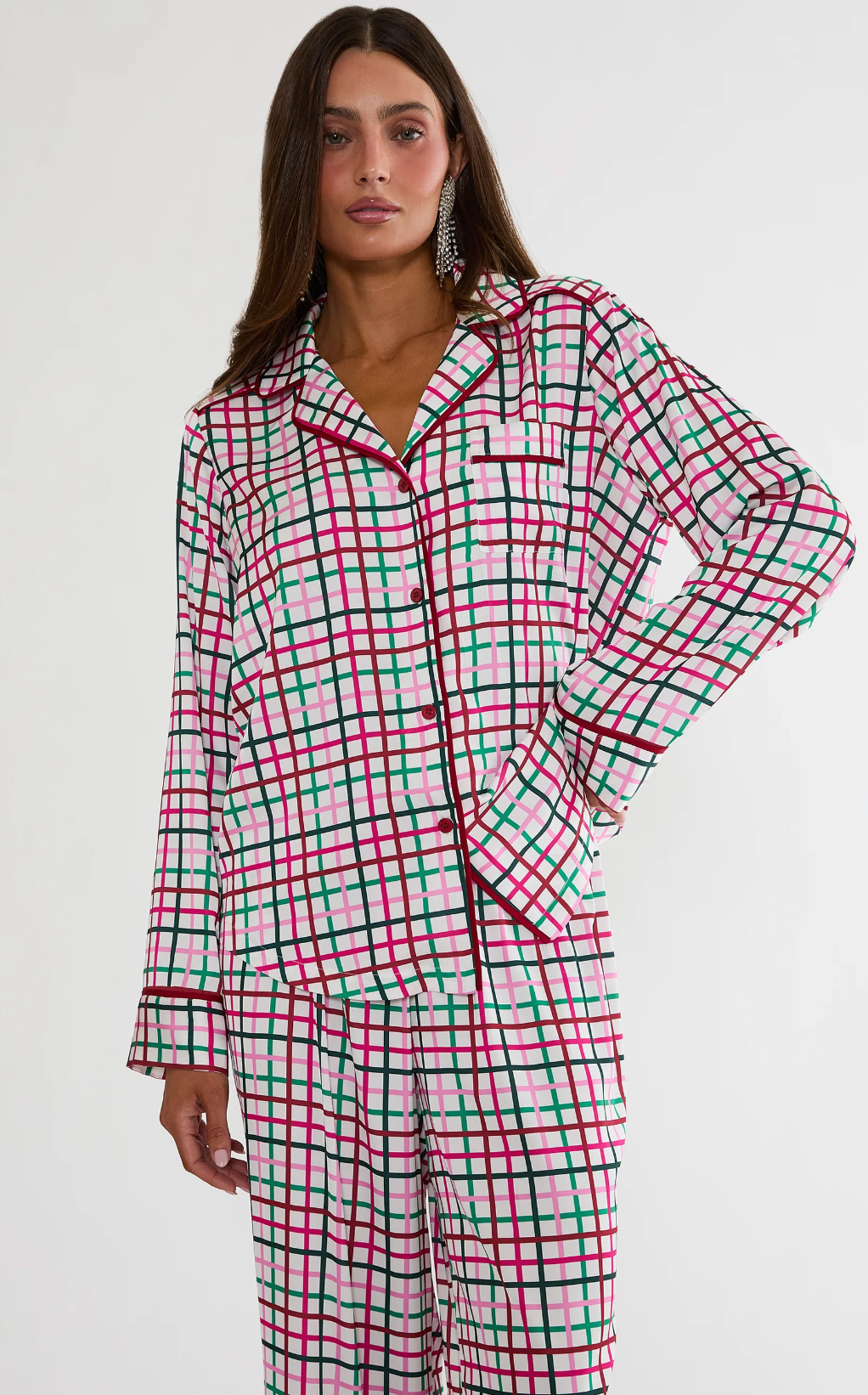 A woman modeling a matching long-sleeve pajama set with a red, green, and black plaid pattern on a white background, photographed against a plain light gray studio backdrop.