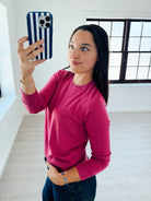 A woman taking a mirror selfie in a bright room, wearing a long-sleeve crewneck knit top in a vibrant magenta pink and dark blue jeans.