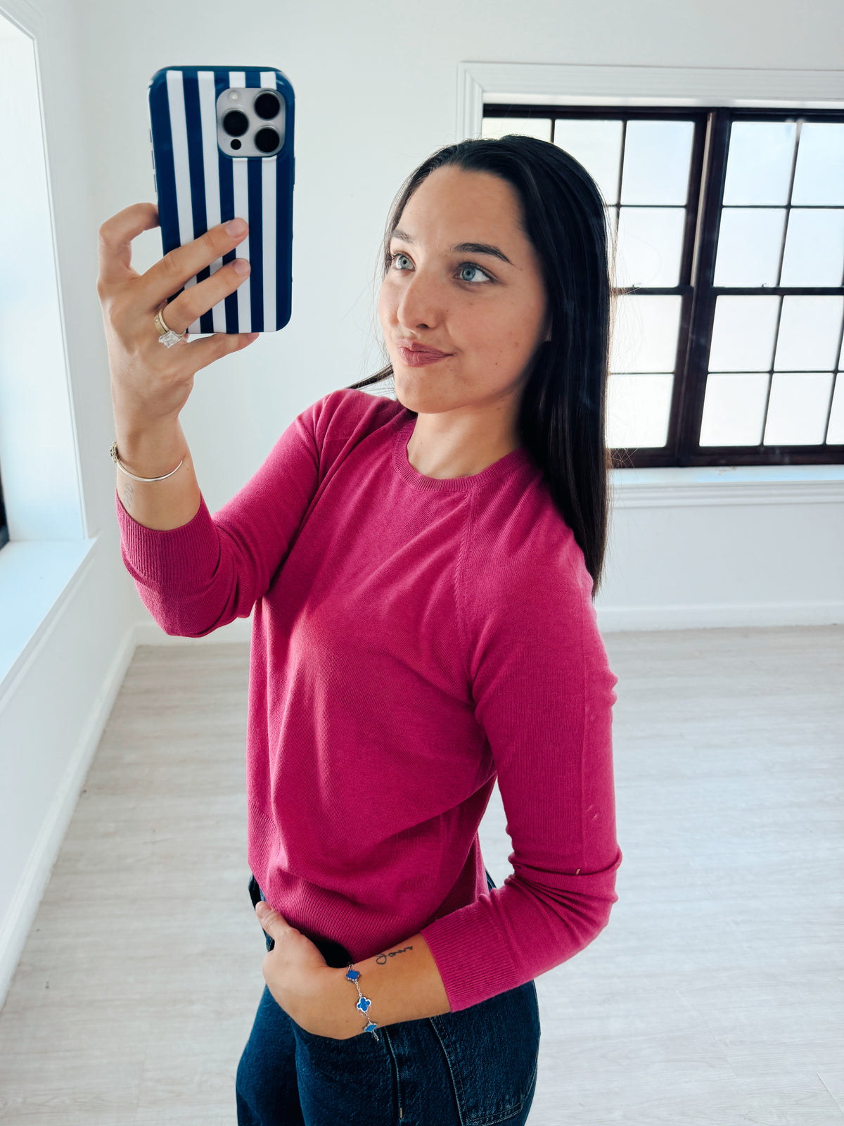 A woman taking a mirror selfie in a bright room, wearing a long-sleeve crewneck knit top in a vibrant magenta pink and dark blue jeans.