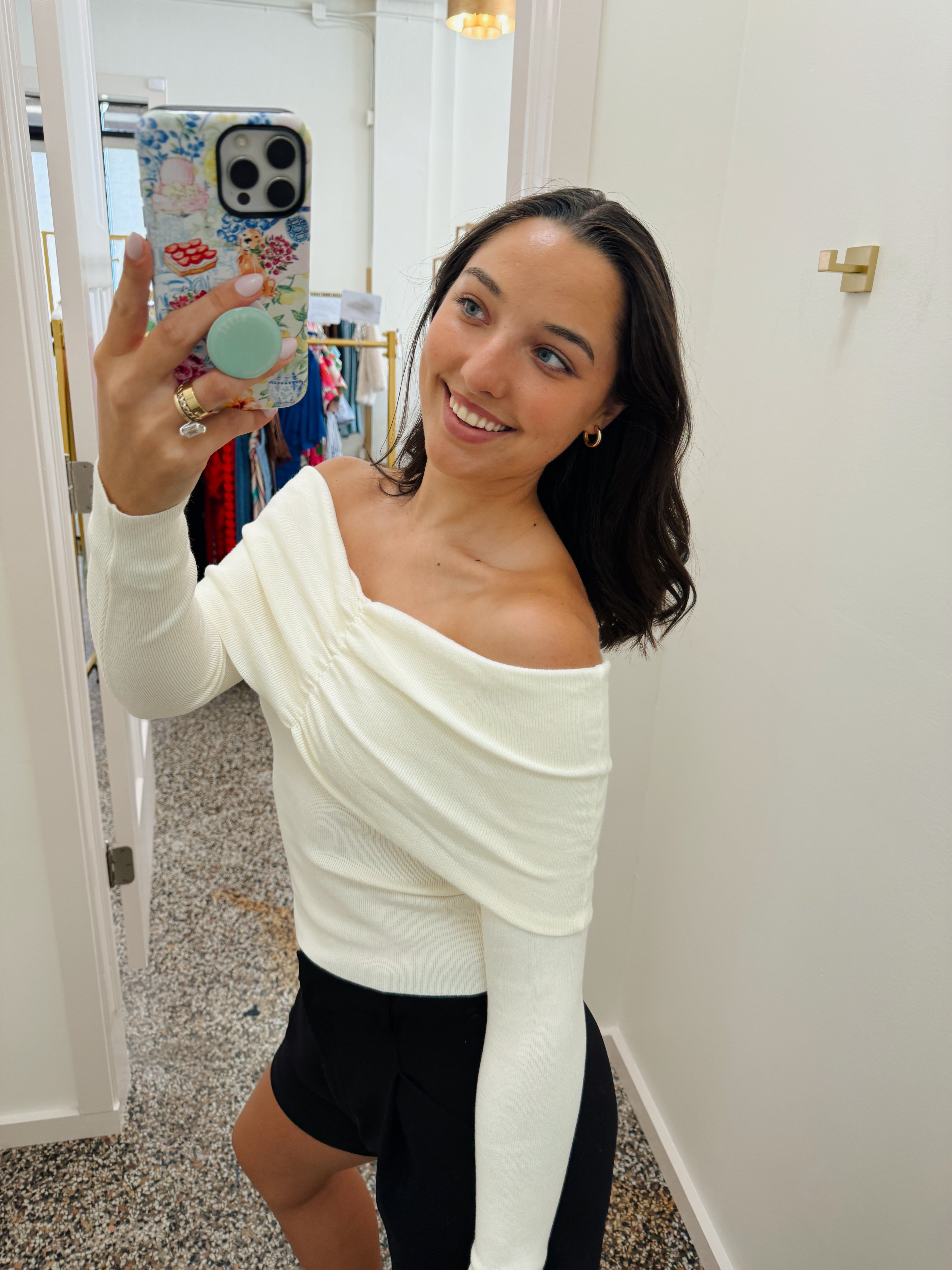 A woman taking a mirror selfie in a fitting room wearing a cream off-the-shoulder long-sleeve top and black shorts.