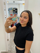 A woman taking a mirror selfie in a boutique, wearing a black short-sleeve crop top and black pants. She is holding a light green circular object in her hand, with a colorful phone case visible in the mirror reflection.