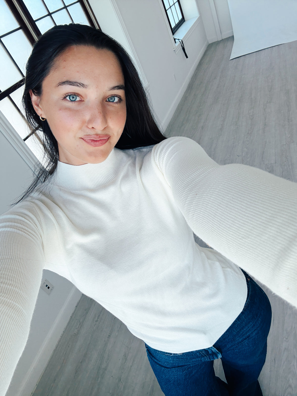 A woman taking a selfie in a bright room, wearing a white ribbed long-sleeve mock neck top and blue jeans.