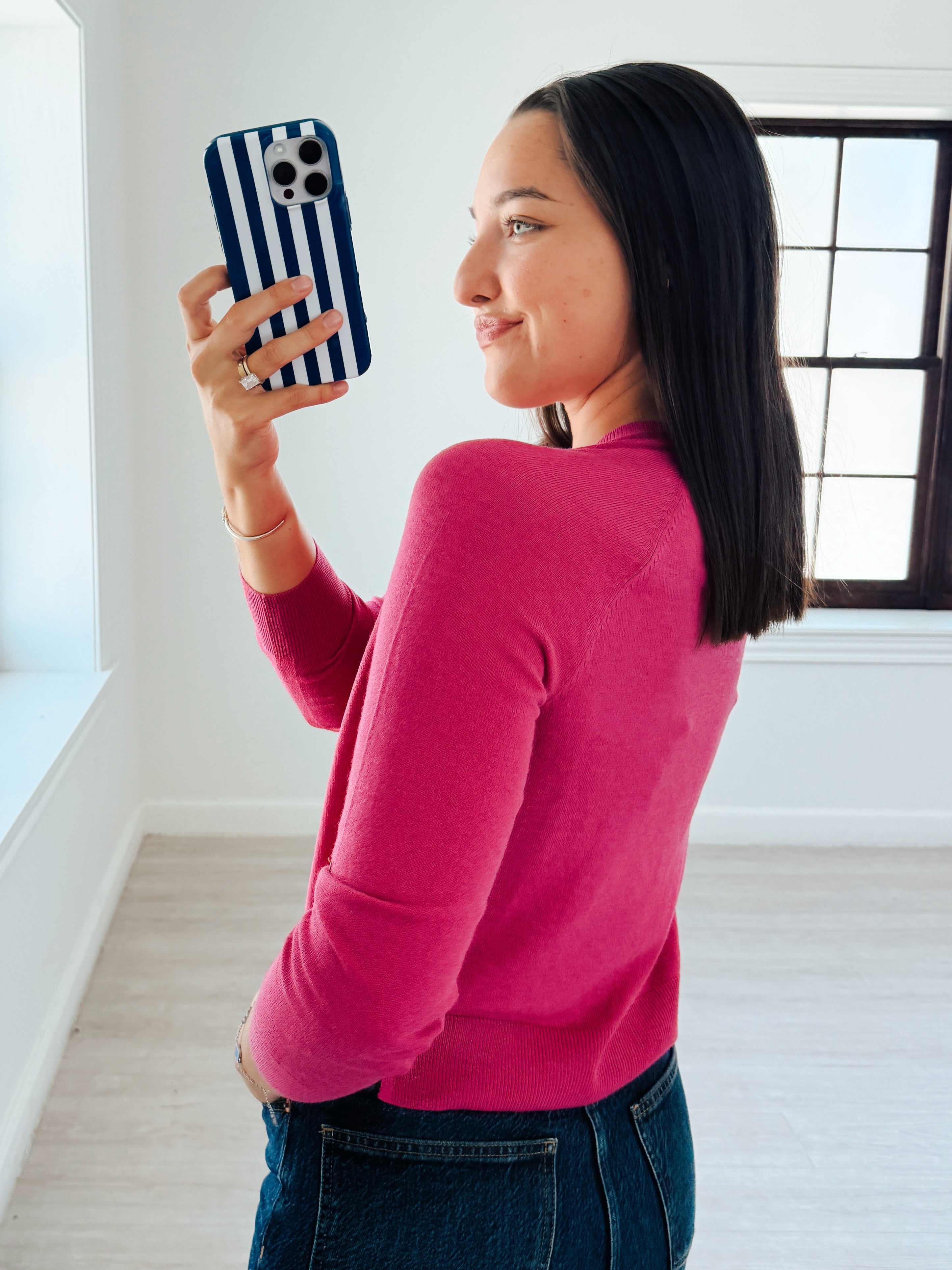 A woman in a bright pink long-sleeve top and blue jeans takes a mirror selfie with a navy and white striped phone case in a bright, modern room.