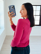 A woman in a bright pink long-sleeve top and blue jeans takes a mirror selfie with a navy and white striped phone case in a bright, modern room.
