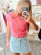 A woman taking a mirror selfie in a boutique, wearing a coral pink short-sleeve button-up top and a light-wash denim mini skirt.