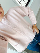 A person wearing a light pink knit cardigan sweater with front buttons and patch pockets, paired with blue jeans in an indoor setting.