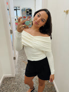 A woman taking a mirror selfie wearing a cream off-the-shoulder long-sleeve top and black shorts in a hallway.