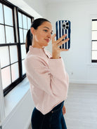 A woman takes a mirror selfie in a bright, modern room with white walls and large black-framed windows. She is wearing a light pink ribbed knit mock-neck sweater and dark blue jeans, holding a blue and white striped phone case.