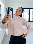 A woman taking a mirror selfie wearing a light pink knit button-up cardigan and dark blue jeans, holding a phone with a navy and white striped case in a bright room with large black-framed windows.