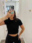 A woman taking a mirror selfie wearing a fitted black short-sleeve crop top and black shorts, with a patterned phone case and bracelets visible.