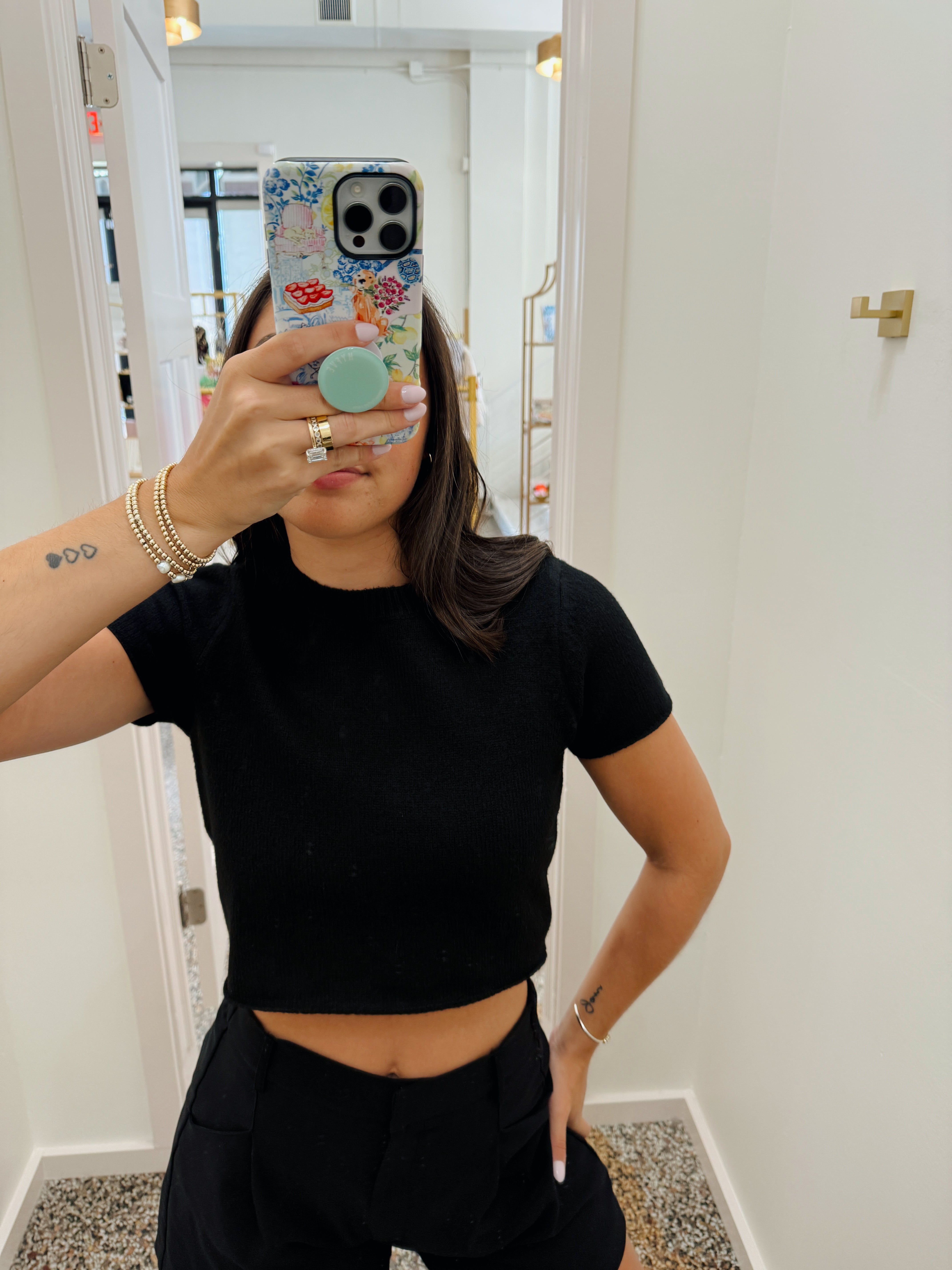 A woman taking a mirror selfie wearing a fitted black short-sleeve crop top and black shorts, with a patterned phone case and bracelets visible.