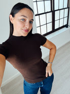 A woman wearing a fitted short-sleeve brown ribbed knit top and blue jeans, taking a mirror selfie in a bright room with a large window.