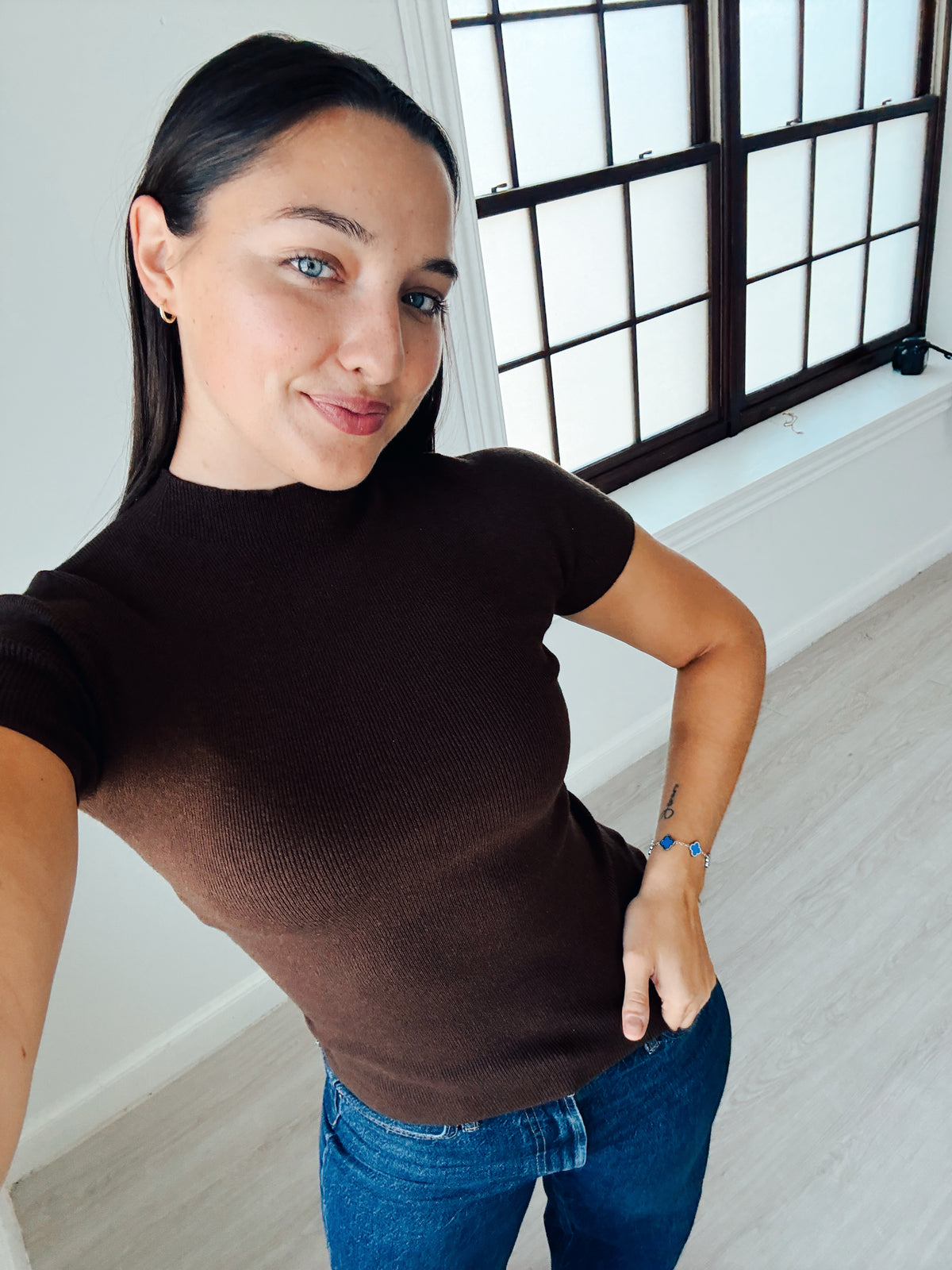 A woman wearing a fitted short-sleeve brown ribbed knit top and blue jeans, taking a mirror selfie in a bright room with a large window.