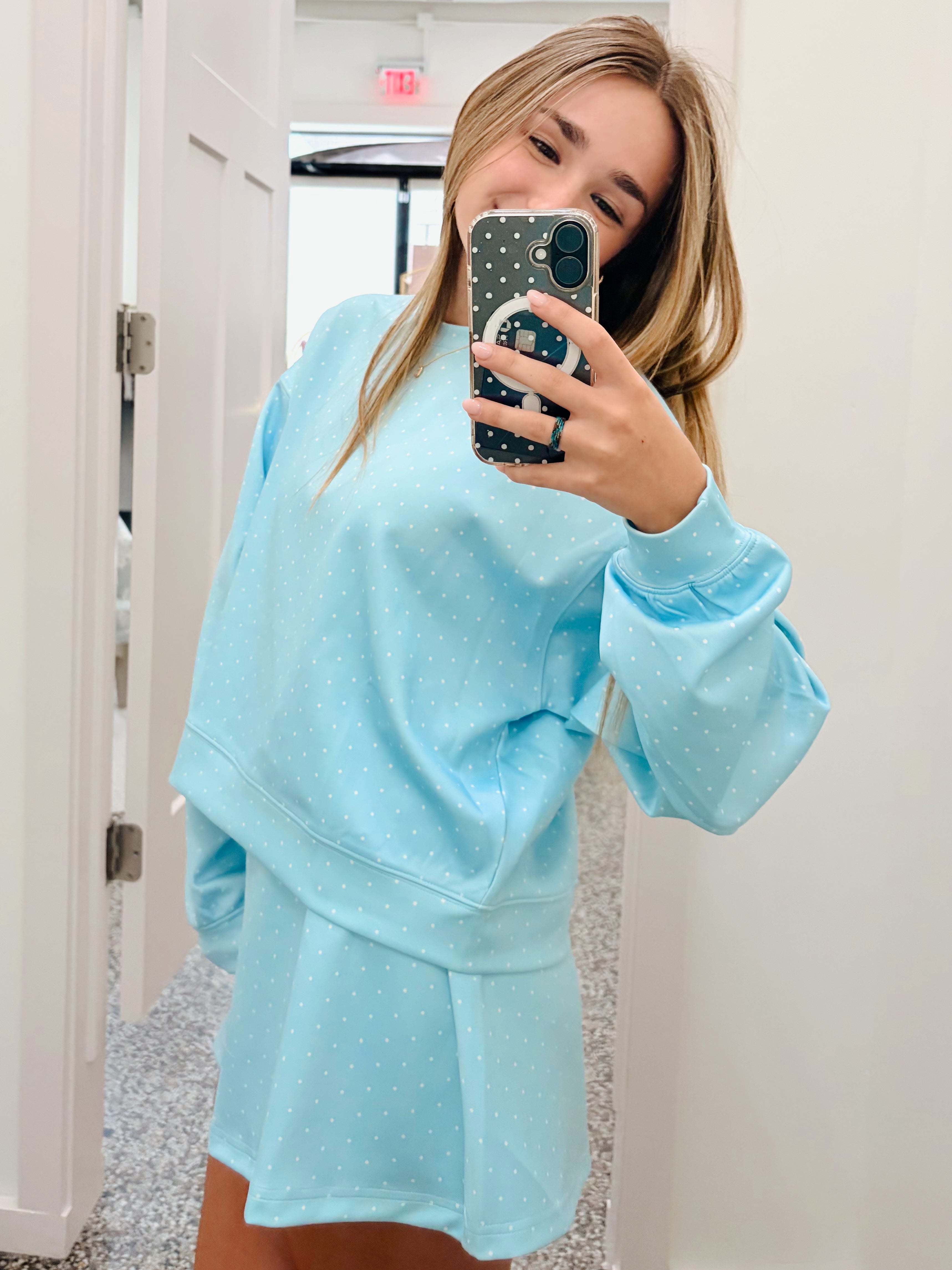 A woman takes a mirror selfie wearing a light blue matching lounge set with small white polka dots, consisting of a sweatshirt and shorts, in a hallway.