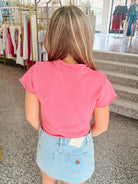 A woman viewed from behind wearing a bright pink short-sleeve ribbed t-shirt and a light blue denim mini skirt inside a clothing store.