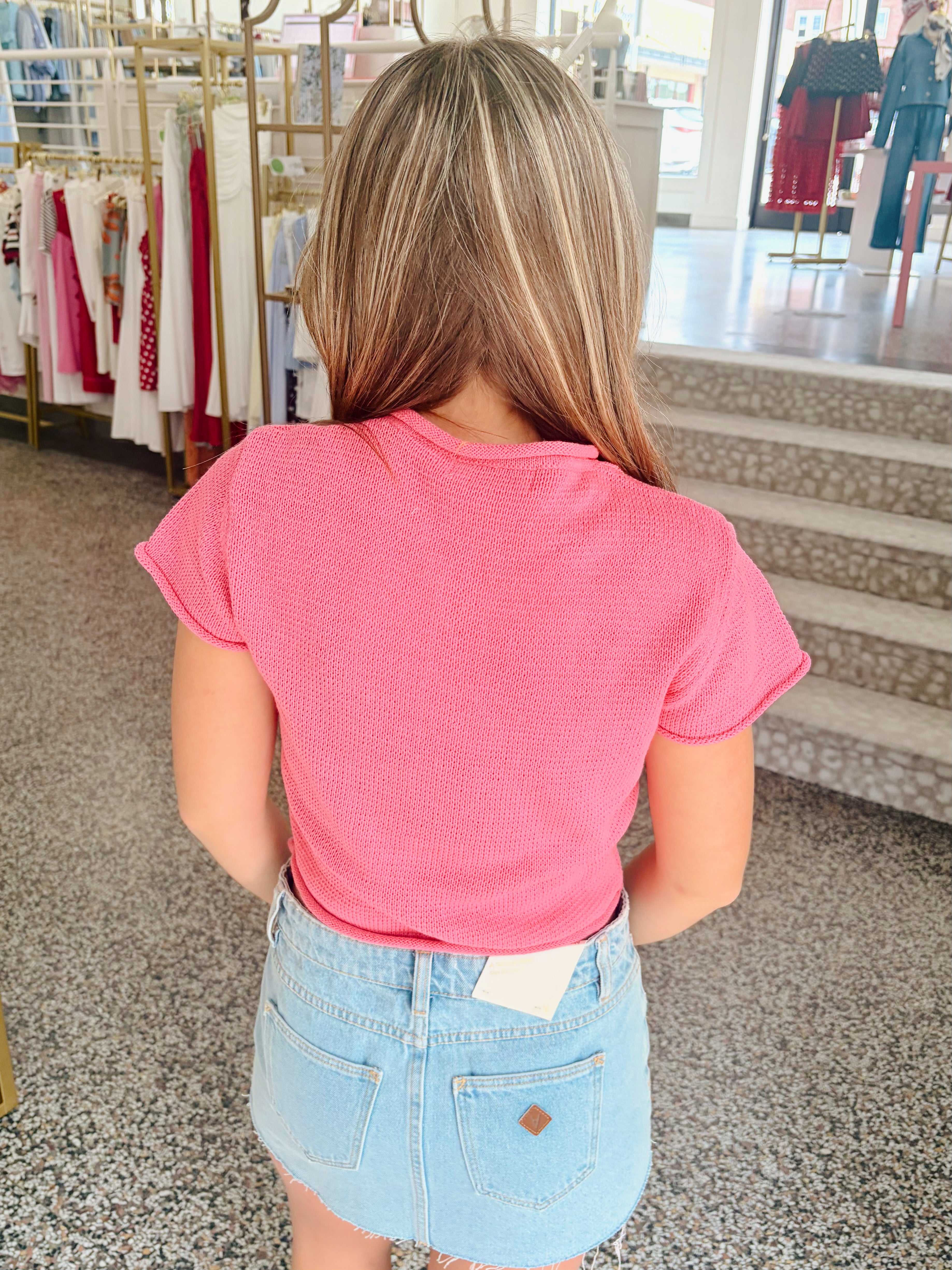 A woman viewed from behind wearing a bright pink short-sleeve ribbed t-shirt and a light blue denim mini skirt inside a clothing store.