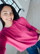A smiling woman wearing a bright pink long-sleeve crewneck sweatshirt and blue jeans, taking a selfie in a modern room with light gray walls and a window.