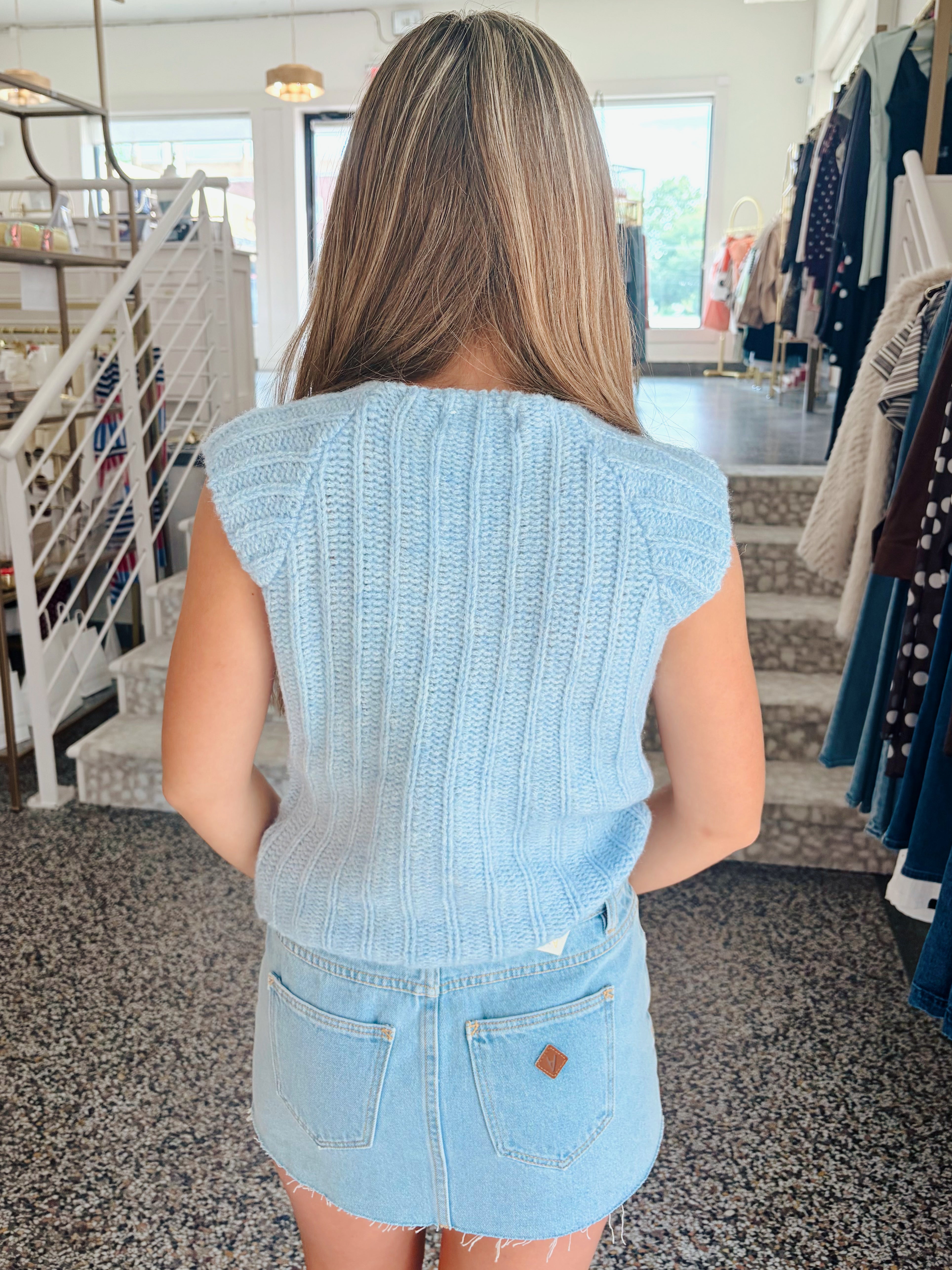 A woman viewed from behind wearing a light blue ribbed knit sleeveless top and a light blue denim mini skirt inside a clothing store.