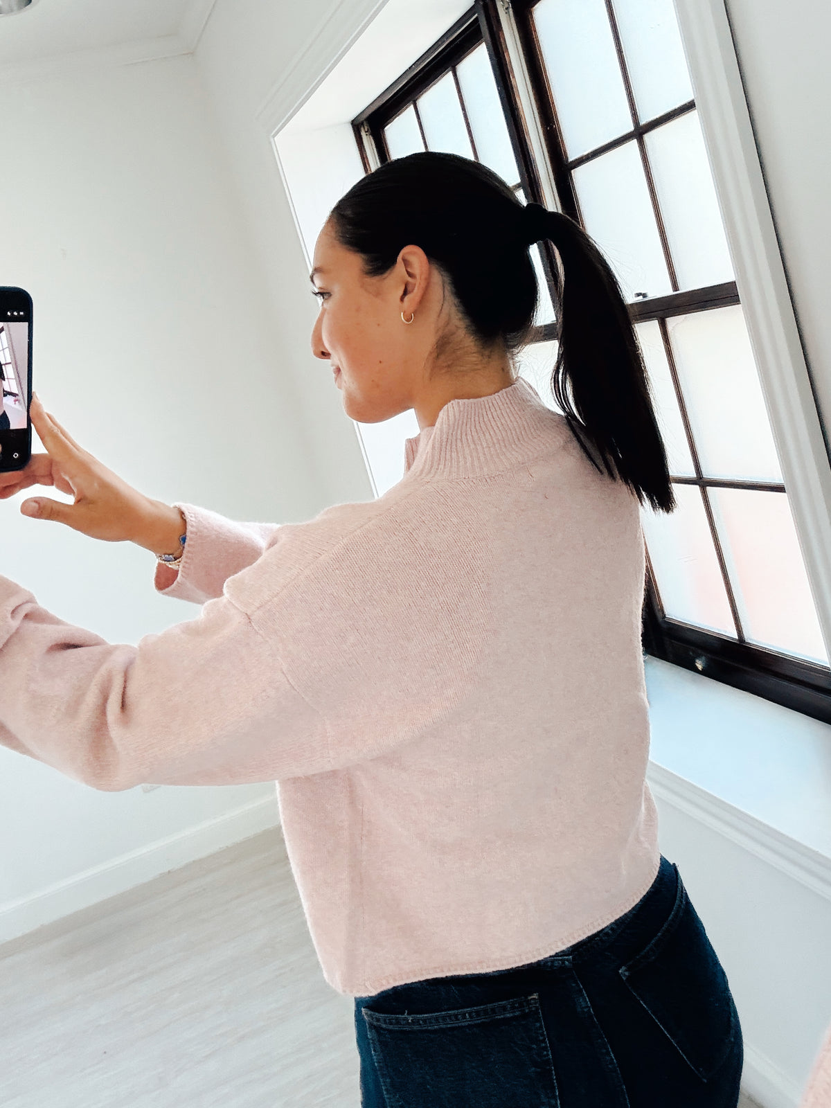 A woman wearing a light pink knit mock neck sweater and dark blue jeans takes a mirror selfie in a bright room with white walls and a black-framed window.
