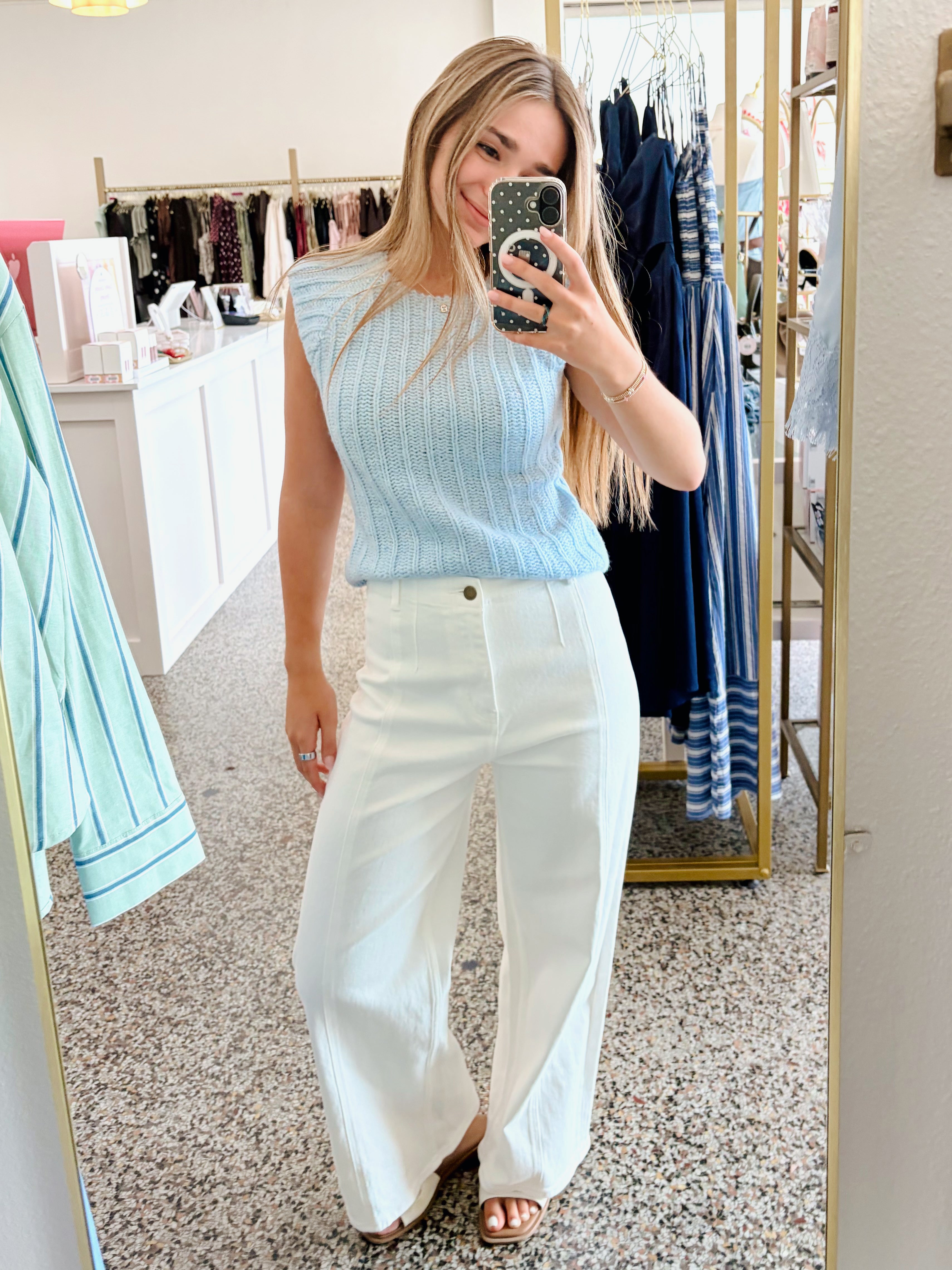 A woman takes a mirror selfie in a boutique wearing a light blue ribbed sleeveless top and white wide-leg pants.