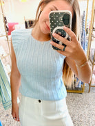 A woman takes a mirror selfie in a boutique while wearing a light blue ribbed knit sleeveless top and white pants. She is holding a black polka-dot phone case.