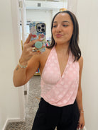 A woman taking a mirror selfie in a fitting room wearing a light pink wrap-style halter top with white polka dots and black pants.