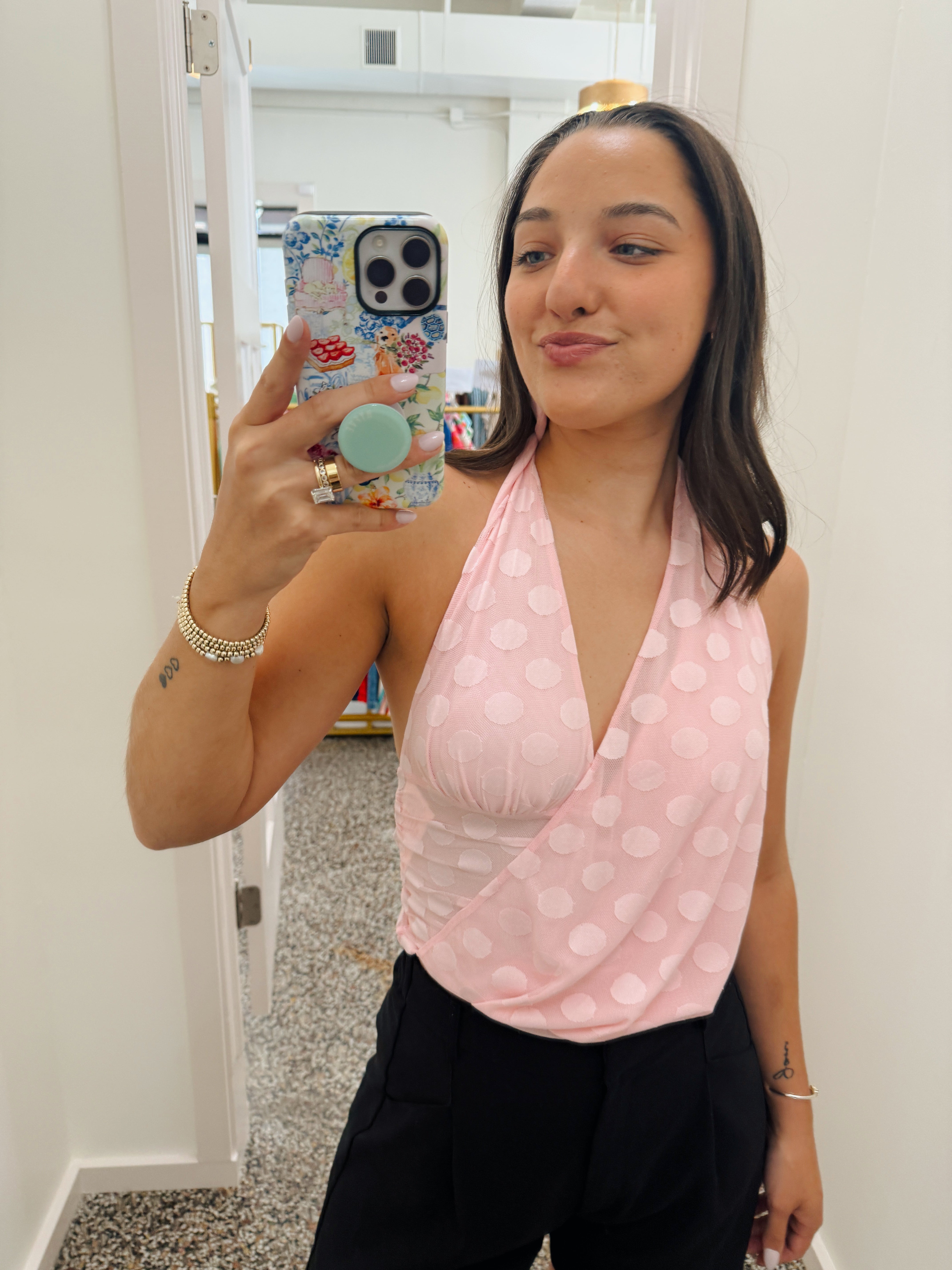 A woman taking a mirror selfie in a fitting room wearing a light pink wrap-style halter top with white polka dots and black pants.