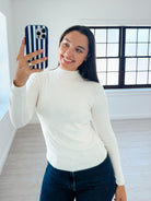 A woman taking a mirror selfie in a bright, modern room, wearing a fitted white ribbed mock neck long-sleeve top and dark blue jeans.