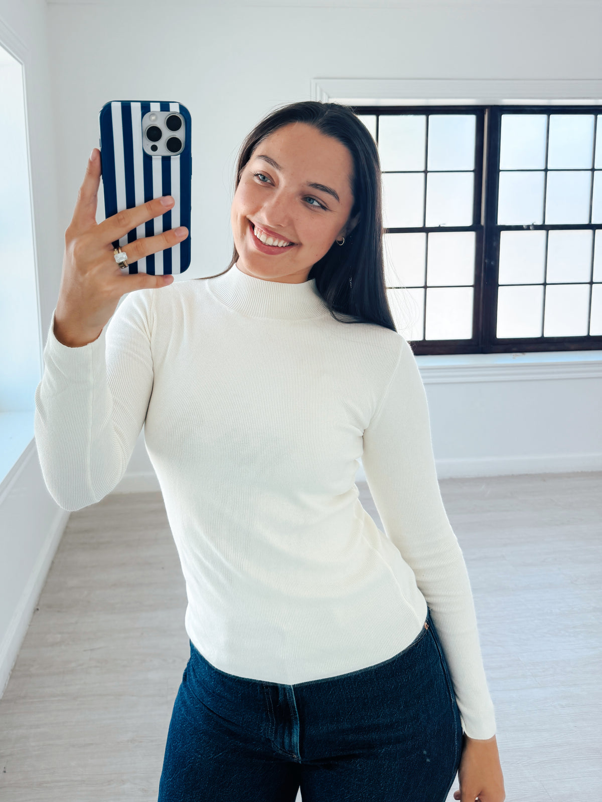A woman taking a mirror selfie in a bright, modern room, wearing a fitted white ribbed mock neck long-sleeve top and dark blue jeans.