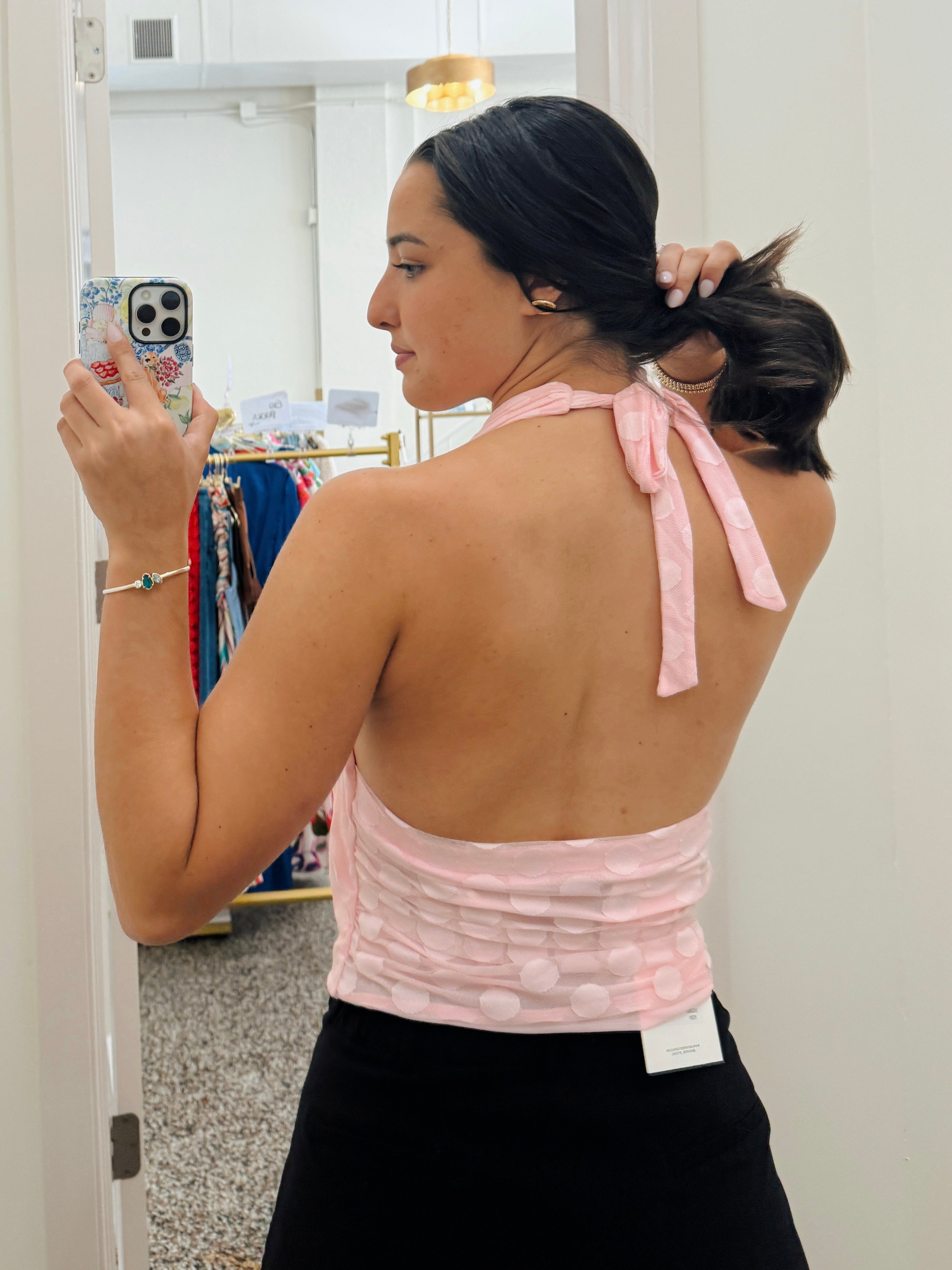 A woman takes a mirror selfie in a fitting room, showing the back of a light pink textured halter top with tie straps and a ruched waistband, paired with a black skirt.