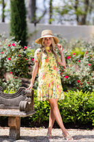 Woman in a yellow floral dress and hat standing in a garden.