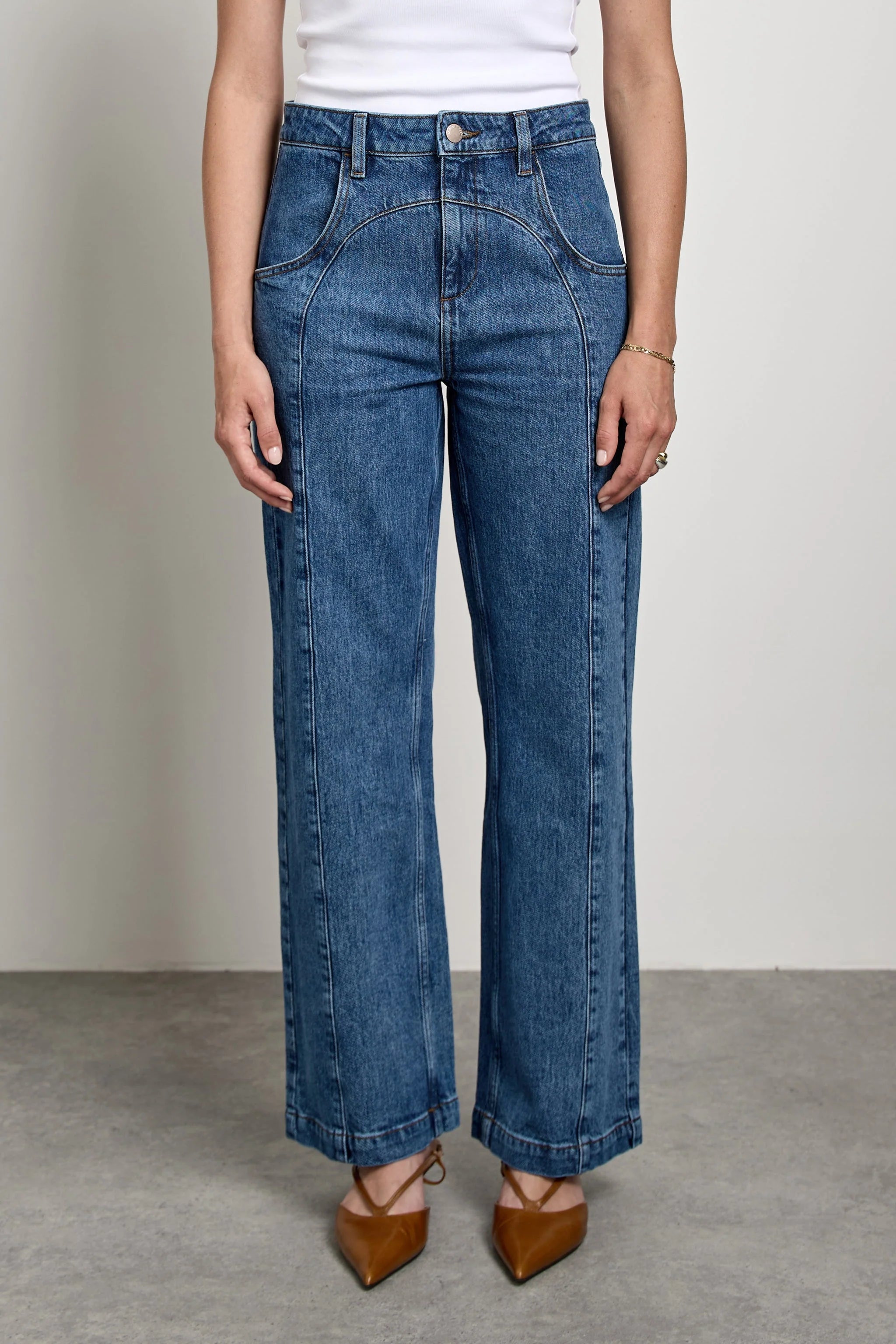 A model wearing a pair of high-waisted, wide-leg blue denim jeans with front pleats, paired with a white top and brown pointed-toe heels, standing against a neutral studio background.