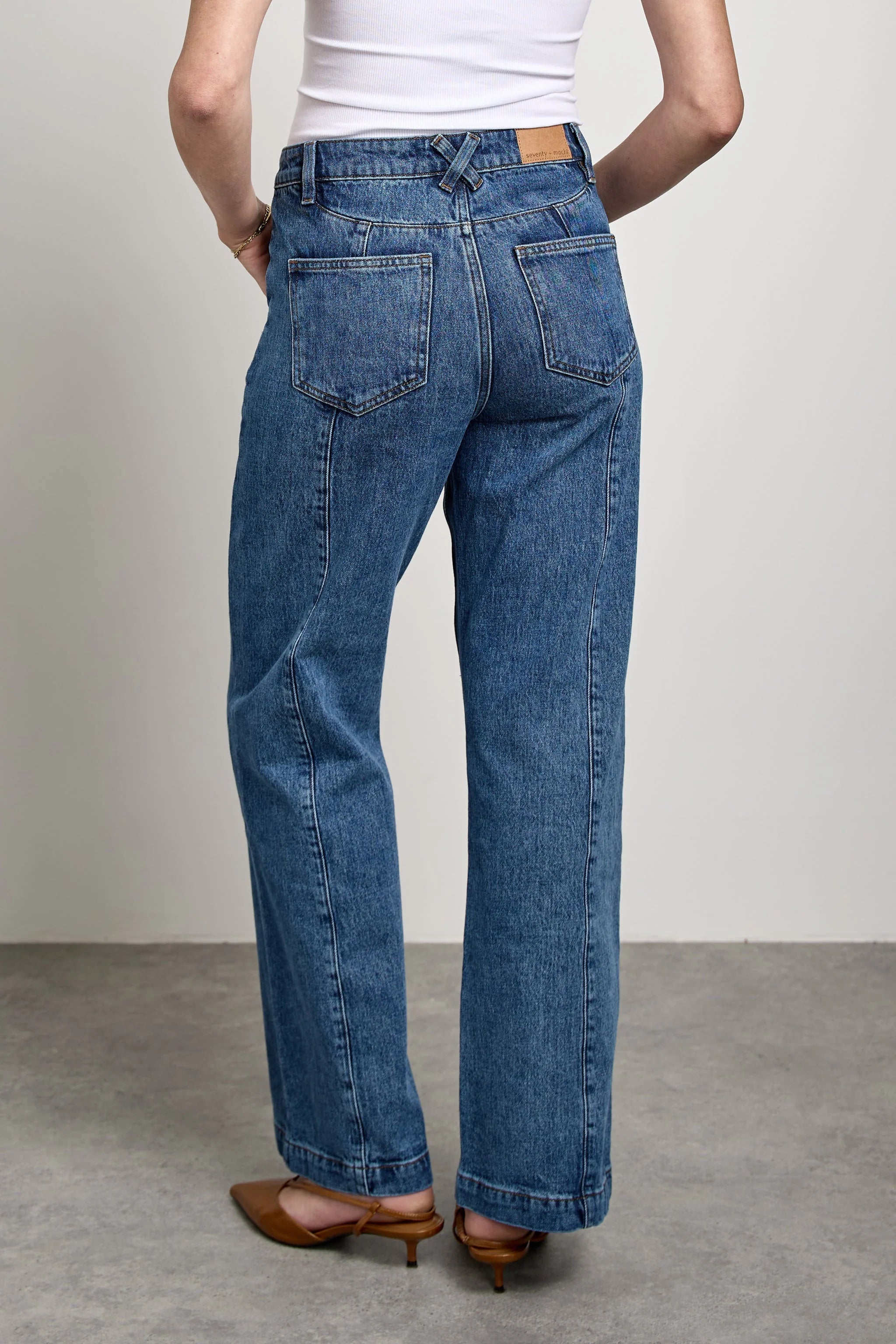 A person shown from the back wearing medium-wash blue straight-leg jeans, a white ribbed top, and brown pointed-toe mule heels against a plain light gray studio background.