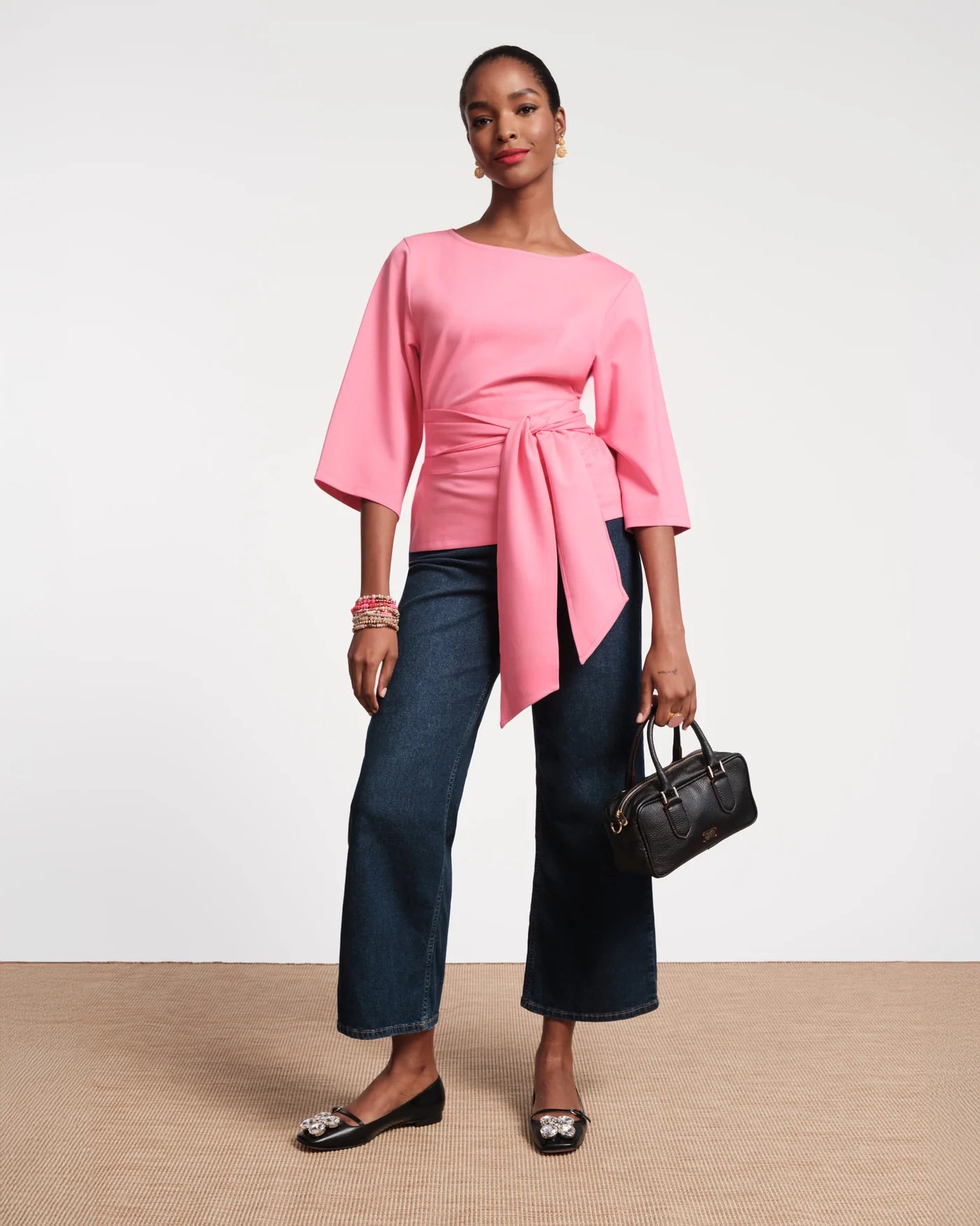 Woman wearing a pink top and blue jeans holding a black handbag on a plain background