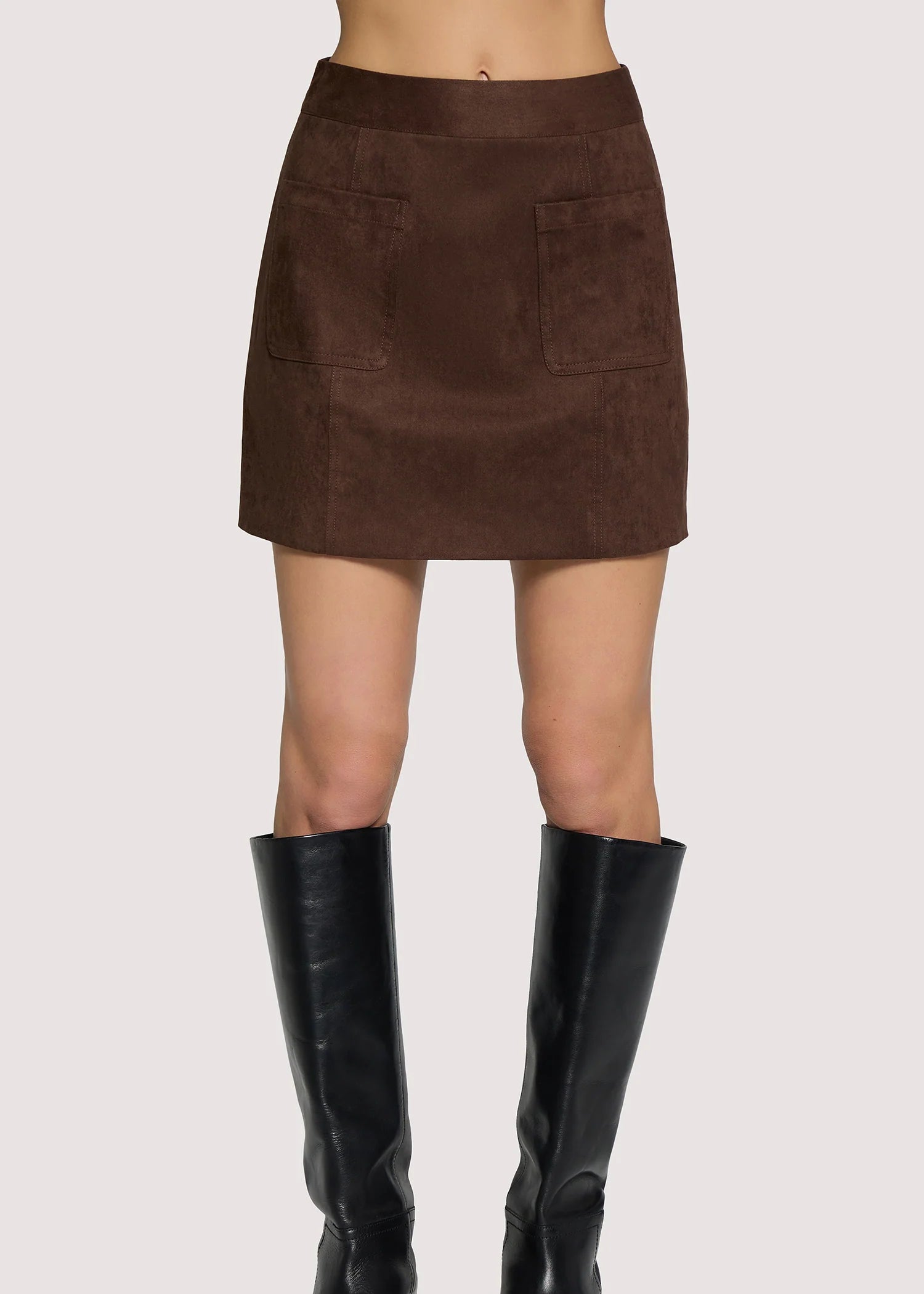 A brown mini skirt with two front patch pockets, shown on a model from the waist down wearing black knee-high leather boots against a light gray background.
