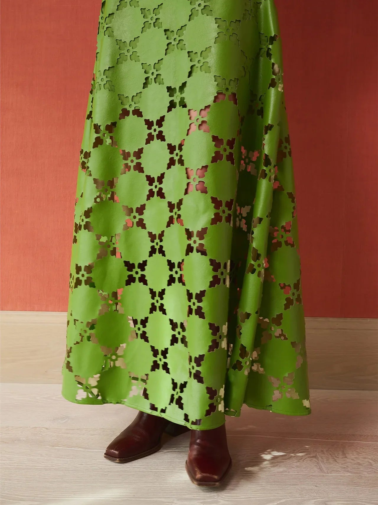 A green maxi skirt with a geometric cutout pattern, shown on a model wearing brown boots against a terracotta-colored wall and light wood floor.