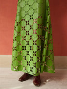 A green maxi skirt with a geometric cutout pattern, shown on a model wearing brown boots against a terracotta-colored wall and light wood floor.