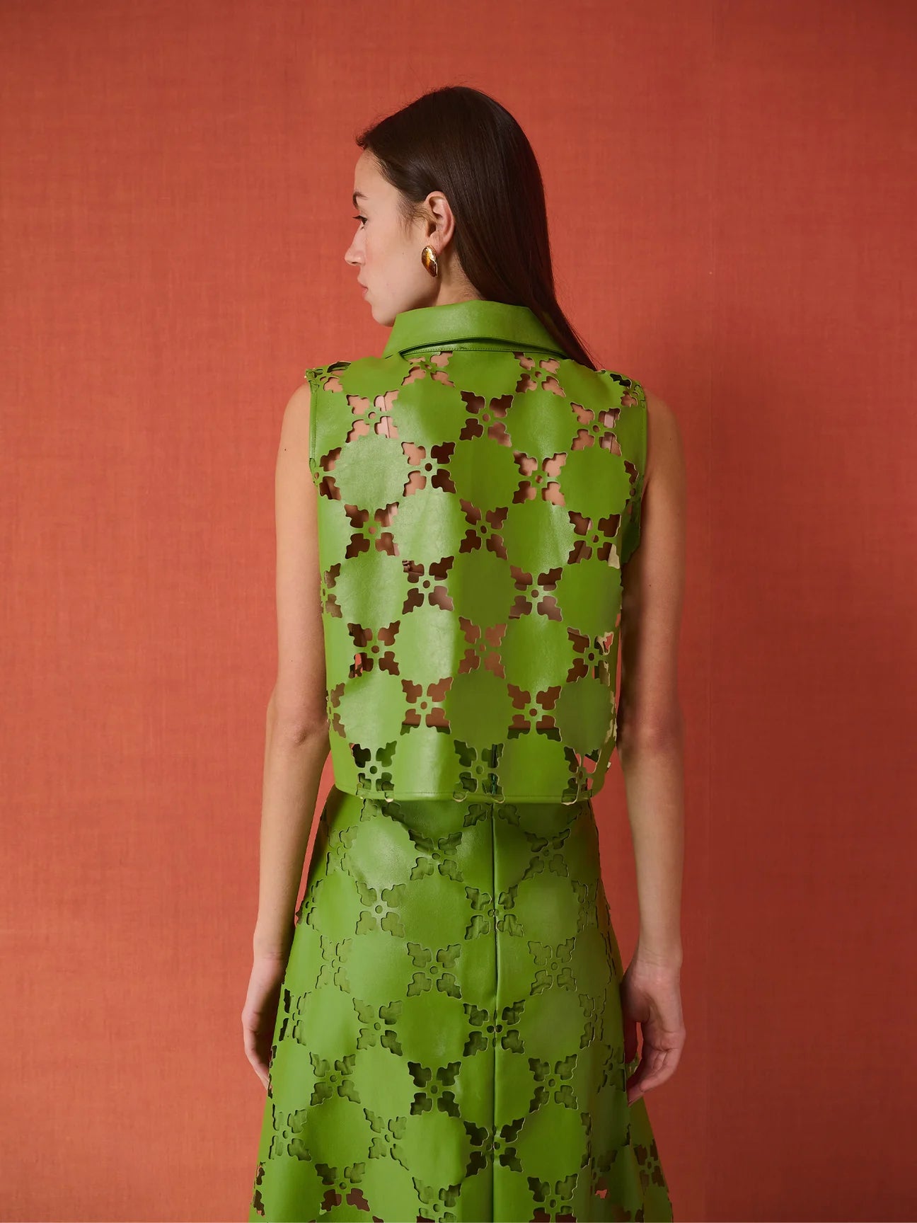 A woman shown from the back wearing a sleeveless green dress with an allover circular cutout pattern, posed against a textured terracotta background.