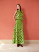 A woman models a sleeveless green two-piece set consisting of a cropped top with a cutout detail and a matching maxi skirt, both featuring a geometric pattern of green circles and brown floral accents, set against a textured orange wall.