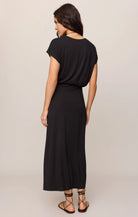 A woman shown from the back wearing a black short-sleeve maxi dress with a draped, loose fit and strappy flat sandals, photographed against a plain light beige studio background.
