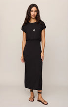 A woman modeling a black midi dress with short sleeves and a gathered waist, paired with brown strappy flat sandals against a light beige studio background.